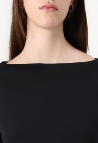 Black top with a wide, flat neckline. Soft, smooth fabric texture. Model's neck is visible, with small gold hoop earrings as an accent.