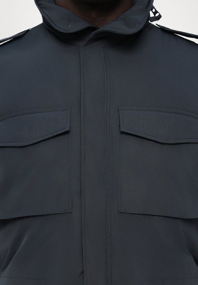 Chaqueta de nailon azul oscuro con cuello alto, dos bolsillos en el pecho y detalles de botones. Textura suave y diseño minimalista.
