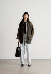 Αδιάβροχο μπουφάν ελιάς με τσέπες, φορεμένο πάνω από μαύρο τοπ. Ανοιχτό μπλε παντελόνι και μαύρη τσάντα. Προσιτά παπούτσια, μαύρο καπέλο-κουβάς.