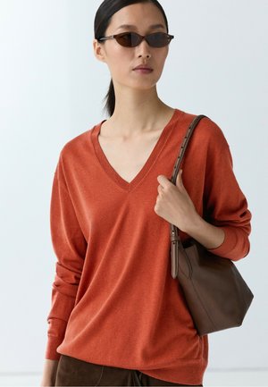 Donna con occhiali da sole scuri, maglione arancione a scollo a V, che tiene una tracolla di borsa marrone sulla spalla, con i capelli raccolti.