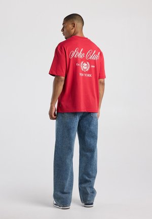 Mann trägt rotes Soho Club T-Shirt und blaue Jeans, steht mit dem Rücken zu einem hellen einfarbigen Hintergrund.
