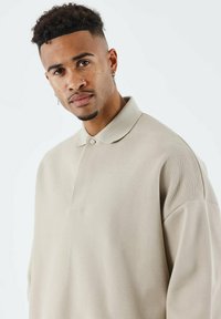 Polo beige con colletto arrotondato, chiusura a bottoni e tessuto lavorato; presenta spalle scese e una vestibilità comoda.