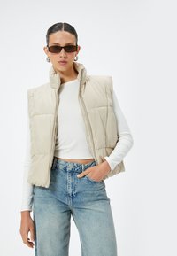 Beige Daunenweste mit hohem Kragen und Reißverschluss, getragen über einem weißen Langarmoberteil. Blaue hochgeschnittene Jeans aus Denim, mit minimalem Schmuck.