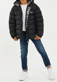 Chaqueta acolchada negra con capucha, que presenta un logo blanco en el lateral y bolsillos con cremallera, combinada con jeans azules y zapatillas blancas.