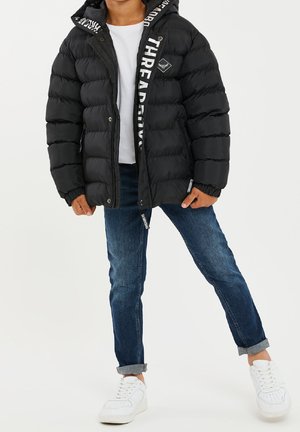 Černá bunda s péřovým vzhledem a kapucí, s bílým logem na boku a kapsami na zip, v kombinaci modrými džíny a bílými teniskami.