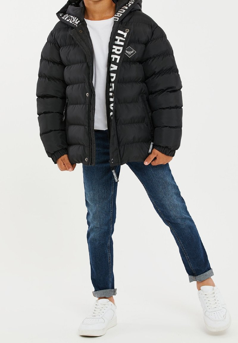Chaqueta acolchada negra con capucha, que presenta un logo blanco en el lateral y bolsillos con cremallera, combinada con jeans azules y zapatillas blancas.