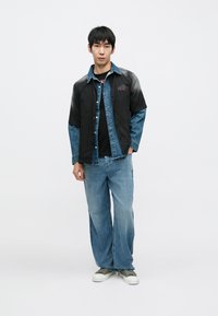 Farmerdzseki fekete és kék részekkel, grafikai részlettel, alatta fekete póló, világoskék bő szárú farmerrel és szürke tornacipővel párosítva.