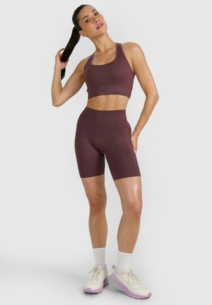 Kobieta w bordowym sportowym biustonoszu i szortach trzymająca kucyk, stojąca z jedną ręką na biodrze, ubrana w białe skarpetki i buty sportowe z różowymi podeszwami.
