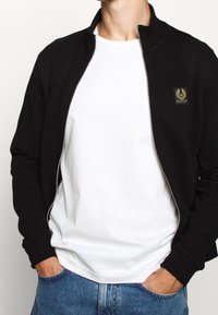 Svart zip-up jacka med krage, tillsammans med en vit t-shirt. Har ribbade muddarna och en liten logotyp patch på vänster sida.