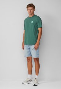 Groen katoenen T-shirt met een ronde halslijn en een klein logo, gecombineerd met lichtblauwe denim shorts met scheuren en zakken. Witte sportschoenen.