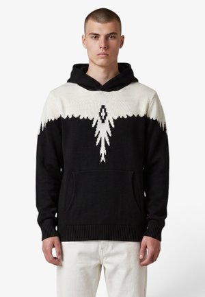 Giovane uomo con indosso una felpa con cappuccio bianca e nera con un motivo astratto bianco sul petto, abbinata a pantaloni bianchi, in piedi su uno sfondo bianco.