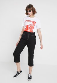 Vit t-shirt med röd grafisk tryck, kombinerad med svarta avklippta denimbyxor och svarta sneakers med vita sulor. Accessoarer inkluderar mörka solglasögon.