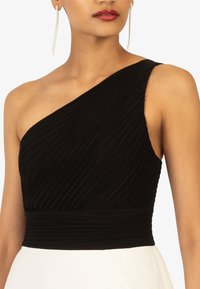 Schwarzes Ein-Schulter-Kleid mit plissierter Textur und tailliertem Oberteil, das einen kontrastierenden weißen Rock und hängende silberne Ohrringe zeigt.