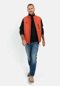 Quilted orange Weste über einem schwarzen gerippten Pullover, blauen Jeans und beigen Stiefeln. Mit Stehkragen und Reißverschlusstaschen.
