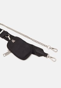 Bolso de cintura de tela negra con cremallera plateada, logo y cadenas de plata desmontables con enganches sobre un fondo blanco.