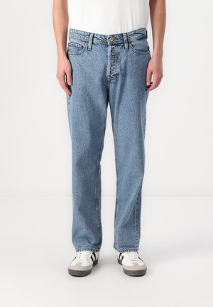 Person in hellblauen, geraden Jeans und weißen Sneakers mit schwarzen Streifen, steht vor einem schlichten weißen Hintergrund.