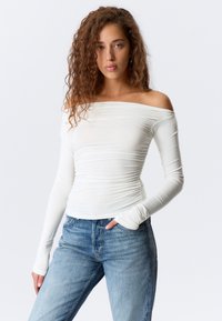 Kvinne med krøllete hår iført en hvit off-shoulder langermet topp og blå jeans, stående med den ene hånden i lommen mot en ensfarget bakgrunn.