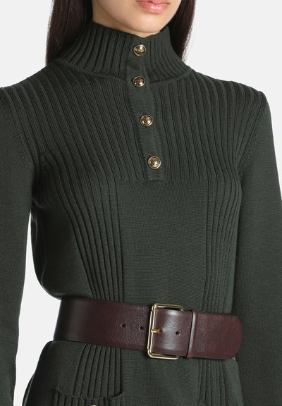Femme portant un pull en maille côtelée vert foncé avec un col montant et des boutons dorés, associé à une large ceinture en cuir marron foncé avec une boucle dorée.
