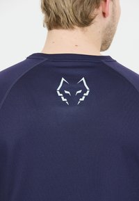 Mand med kort blond hår iført en marineblå skjorte med et stiliseret hvidt ulvehoved-logo på den øverste del af ryggen.