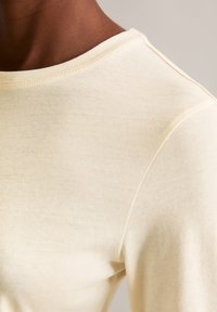 Top de cor creme com uma textura suave e macia, decote arredondado e um discreto detalhe de costura nas costuras dos ombros.