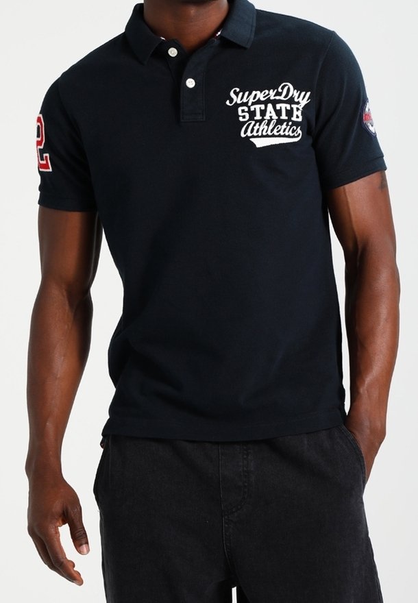 Mann trägt ein schwarzes kurzärmeliges Poloshirt mit dem Schriftzug "SuperDry State Athletics" auf der Brust und der Nummer "2" am Ärmel.