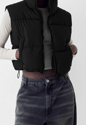 Person wearing a black puffer vest over a light long-sleeve crop top and high-waisted faded black jeans, hands tucked in vest pockets.