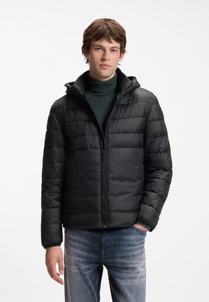 Jeune homme portant une veste matelassée noire zippée sur un pull à col roulé vert foncé et un jean bleu délavé, debout avec une main dans la poche.