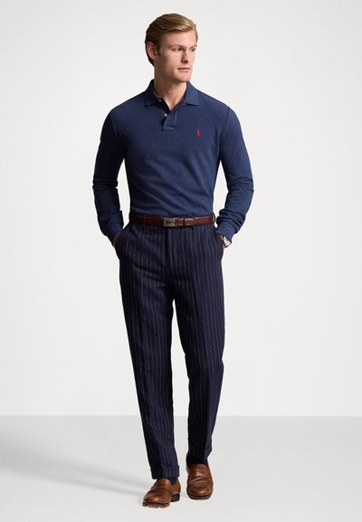 Marineblauwe lange mouw polo, slim-fit marineblauwe broek met streepjes, bruine leren riem en bruine loafers. Subtiele rode logo op de polo.
