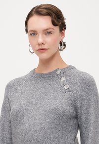 Grå strikket sweater med raglanærmer, prydet med tre dekorative knapper på skulderen. Blød tekstur, tætsiddende design og rund halsudskæring.