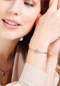 Zilveren armband met een cirkelvormige charm, waarop de gegraveerde letter "S" staat. De vrouw draagt een getextureerde lichtgekleurde top.