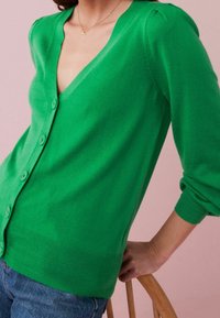 Cardigan verde realizzato in morbido tessuto a maglia, con scollatura a V profonda, chiusura a bottoni e polsini a costine, dal taglio aderente.