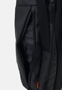 Sac à dos noir en tissu durable; il présente une poche latérale zippée et une doublure texturée, avec un petit accent orange près de la fermeture éclair.