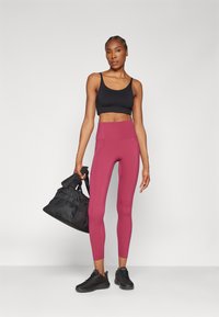 Soutien-gorge de sport noir, leggings bordeaux taille haute, baskets noires, portant un sac de sport noir avec une poche en mesh. Textures lisses, design ajusté.