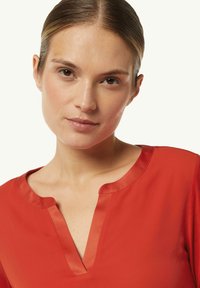 Rote Bluse mit satinierter Oberfläche, V-Ausschnitt und kurzen Ärmeln. Glatte Textur mit einem leicht lockeren Schnitt und dezenten Kragenakzenten.