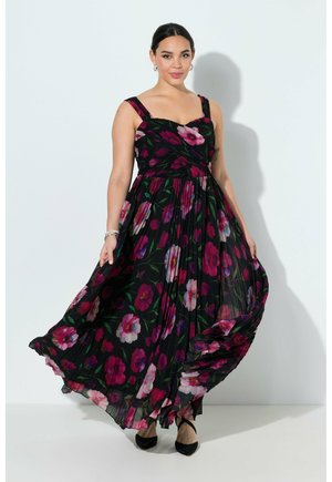 Kvinde iført en flagrende sort maxikjole med pink og lilla blomsterprint, stående mod en ensartet lys baggrund.