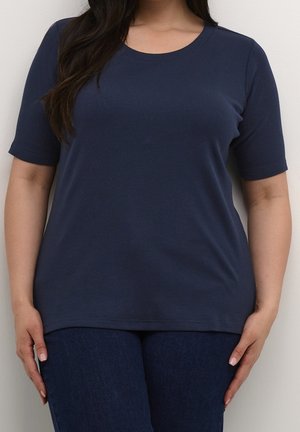 Femme portant un haut bleu marine à manches courtes et col rond, et un jean bleu foncé, debout contre un mur de couleur claire.