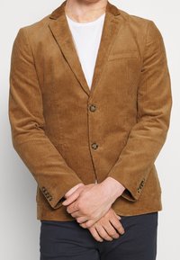 Hnědý kordurový blazer se dvěma knoflíky, klopami a jednou náprsní kapsou. Tkanina má ribbovanou texturu a jemný lesk.