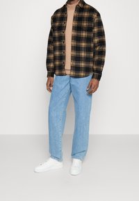 Camicia in flanella a quadri nera e beige sopra una t-shirt beige, abbinata a jeans chiari a gamba dritta e sneakers bianche.