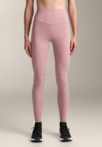 Világos rózsaszín leggings, magas derékkal, rugalmas anyagból készült. Sima textúra, teljes hosszú kialakítás, fekete sportcipővel párosítva.