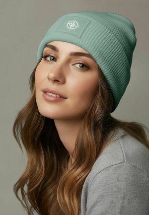 Light green ribbed knit beanie with a rectangular patch featuring a compass design. Styled with long, wavy brown hair and a gray sweater.