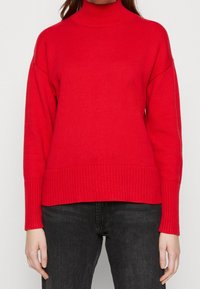 Femme portant un pull à col roulé côtelé rouge à manches longues et un jean noir, se tenant devant un fond blanc uni.