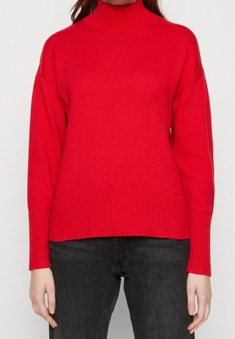 Femme portant un pull à col roulé côtelé rouge à manches longues et un jean noir, se tenant devant un fond blanc uni.