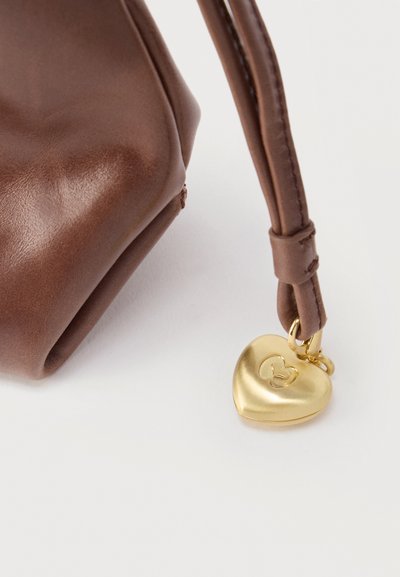 Coin d'un sac en cuir marron avec une sangle attachée présentant un petit charme en forme de cœur doré gravé d'un symbole stylisé.