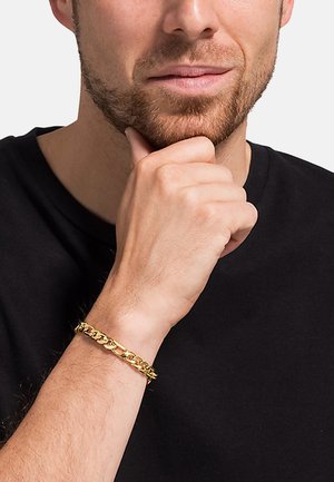 Goldene Kettenarmband an einem Handgelenk, mit verzahnten Gliedern und glänzender Oberfläche, getragen von einer Person, deren Hand auf ihrem Kinn ruht.