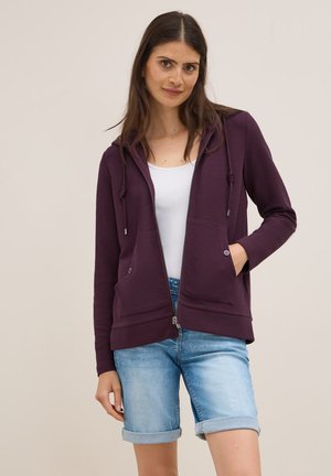 Purple zip-up hoodie with front pockets, drawstring hood, and ribbed hem. Worn over a white shirt and paired with rolled denim shorts.