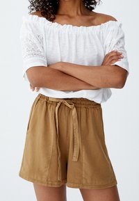 Haut blanc à épaules dénudées avec des accents en dentelle, associé à un short beige avec taille élastique et détail noué. Tissu lisse, coupe décontractée.