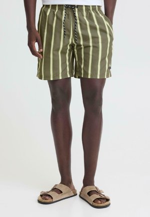 Mand iført olivengrønne shorts med striber, sorte og hvide snore og beige sandaler med spænde, stående på hvidt gulv.