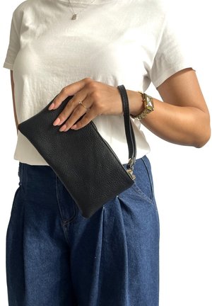 Clutch in pelle nera con finitura textured, dotato di chiusura con zip e di un cinturino per il polso, tenuto in mano su uno sfondo semplice.
