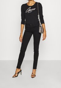 Černý dlouhý rukáv top s třpytivým logem "Guess", spárovaný s černými skinny džíny a černými páskovými podpatky. Stříbrná řetízková kabelka dodává detail.