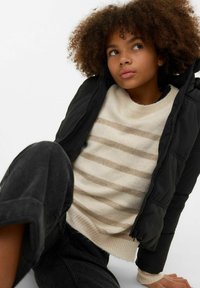 Sort dunjakke med hette, brukt over en kremfarget genser med beige striper, kombinert med mørke denimbukser.